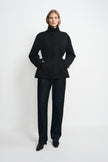 A model wearing a black jacket fastened to the high collar, with a cinched waist and hem flaring below it, paired with dark jeans and pointed black shoes.