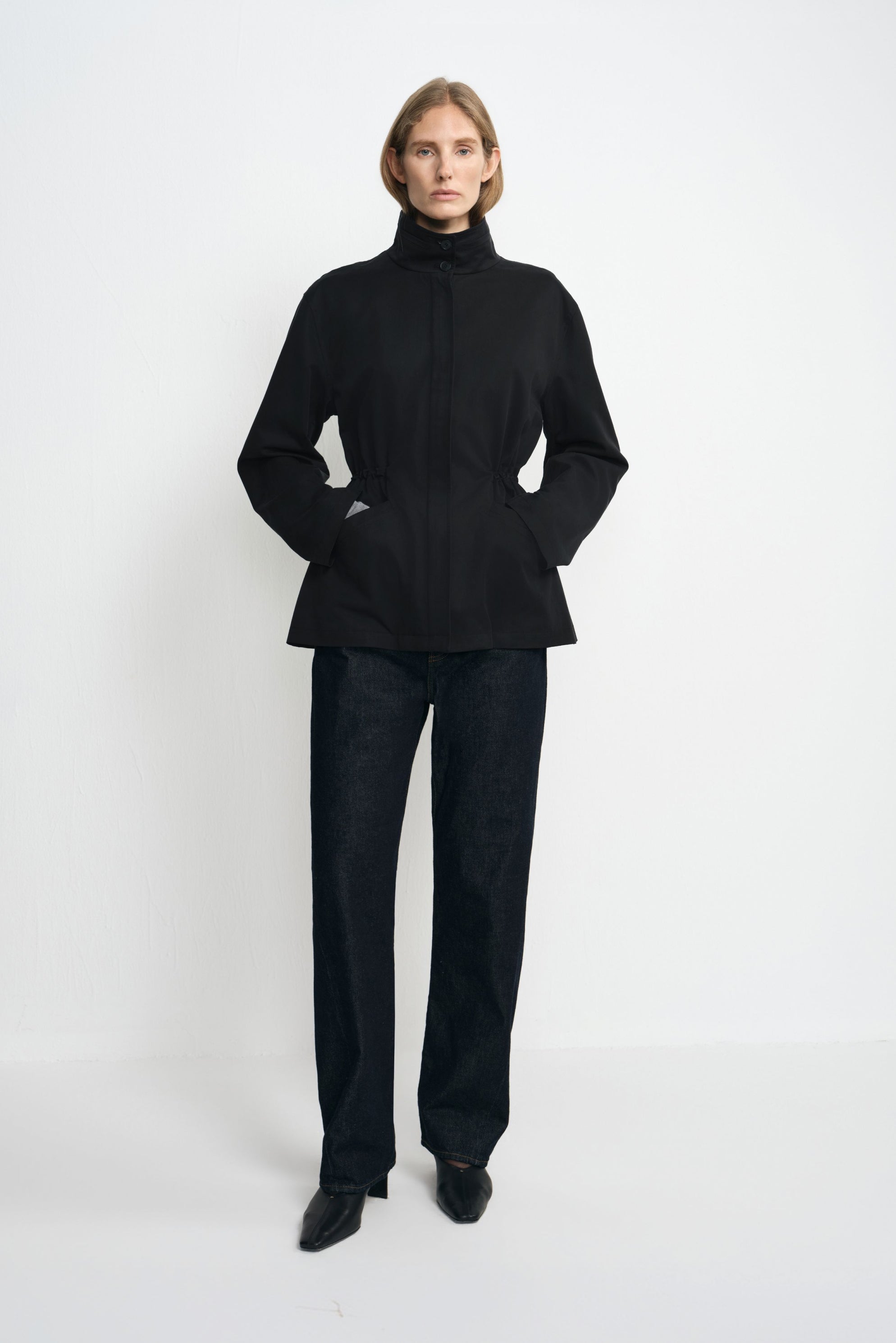 A model wearing a black jacket fastened to the high collar, with a cinched waist and hem flaring below it, paired with dark jeans and pointed black shoes.