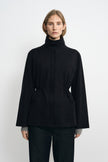 Front detail of a black jacket on a model, showing the two-button stand collar, gathered waist and broad sleeves, with grey cuffs visible underneath.