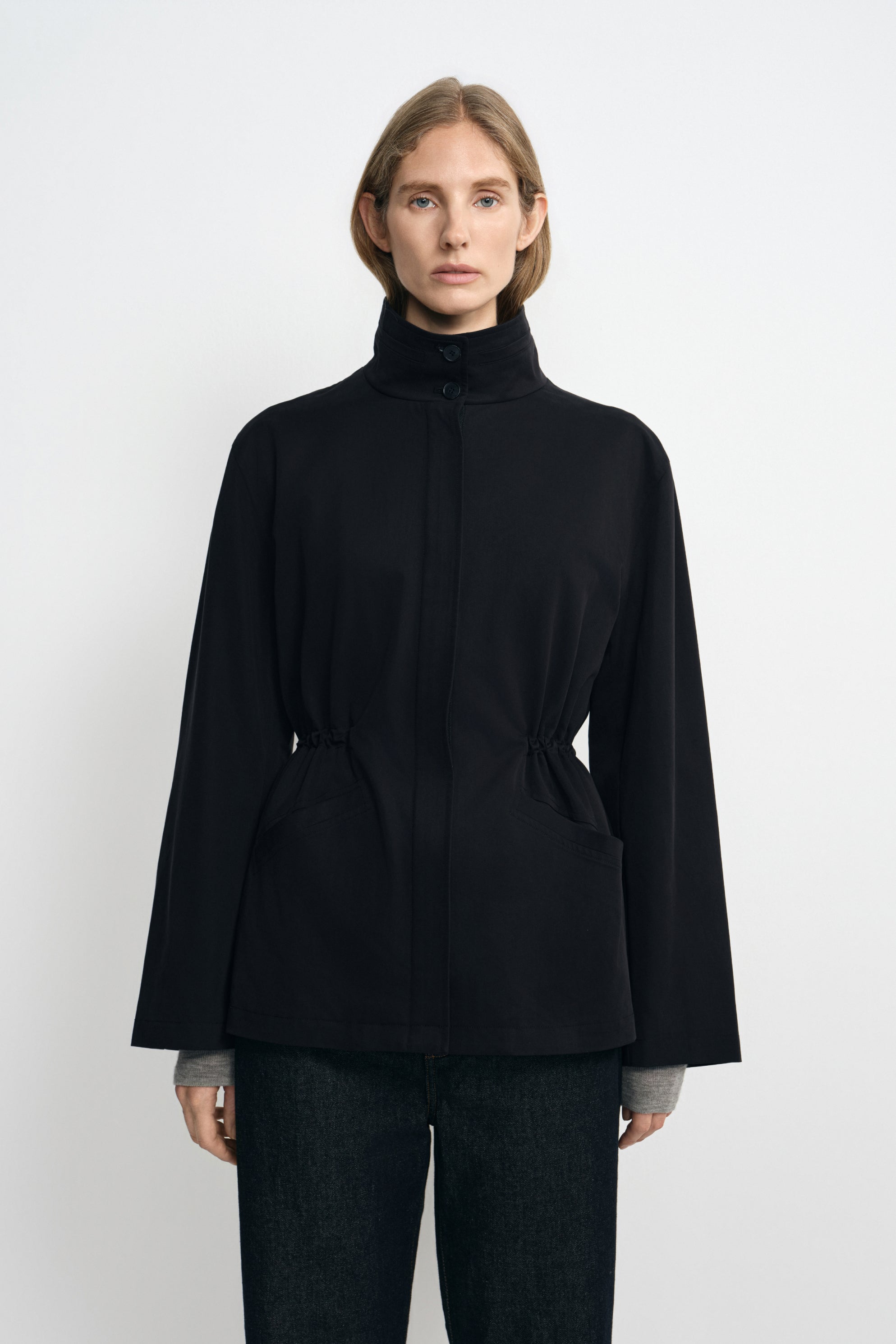 Front detail of a black jacket on a model, showing the two-button stand collar, gathered waist and broad sleeves, with grey cuffs visible underneath.