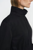 Close-up of a black jacket on a model, showing the tall collar secured with two buttons, parallel stitching around the neck and dropped shoulder seam.