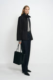 Side view of a model in an open black stand-collar jacket with a loose straight hem, paired with grey layers, dark jeans, black shoes and a black tote.