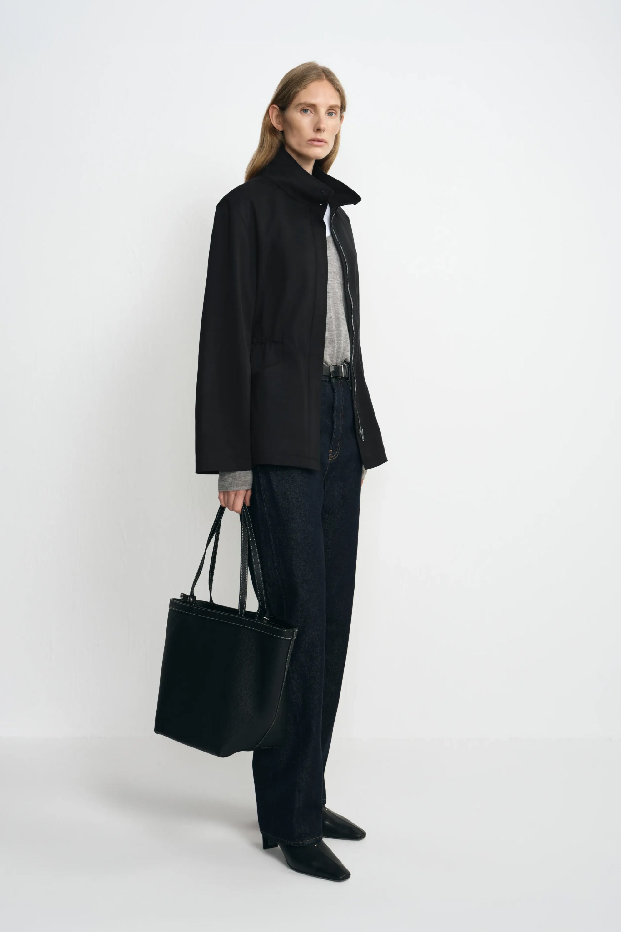 Side view of a model in an open black stand-collar jacket with a loose straight hem, paired with grey layers, dark jeans, black shoes and a black tote.