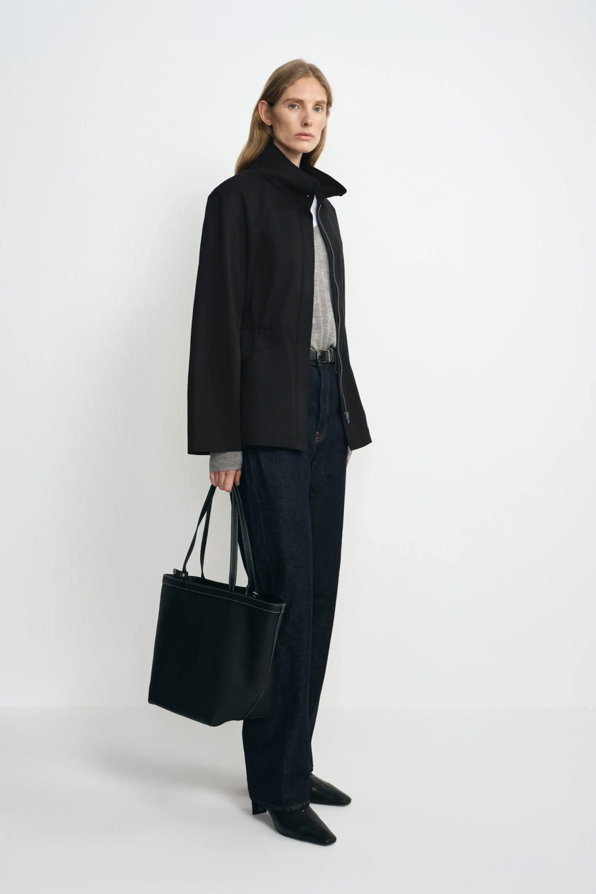 Side view of a model in an open black stand-collar jacket with a loose straight hem, paired with grey layers, dark jeans, black shoes and a black tote.