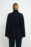 Back view of a model wearing a black jacket, showing the high collar, gathered waist and hem flaring over dark jeans.