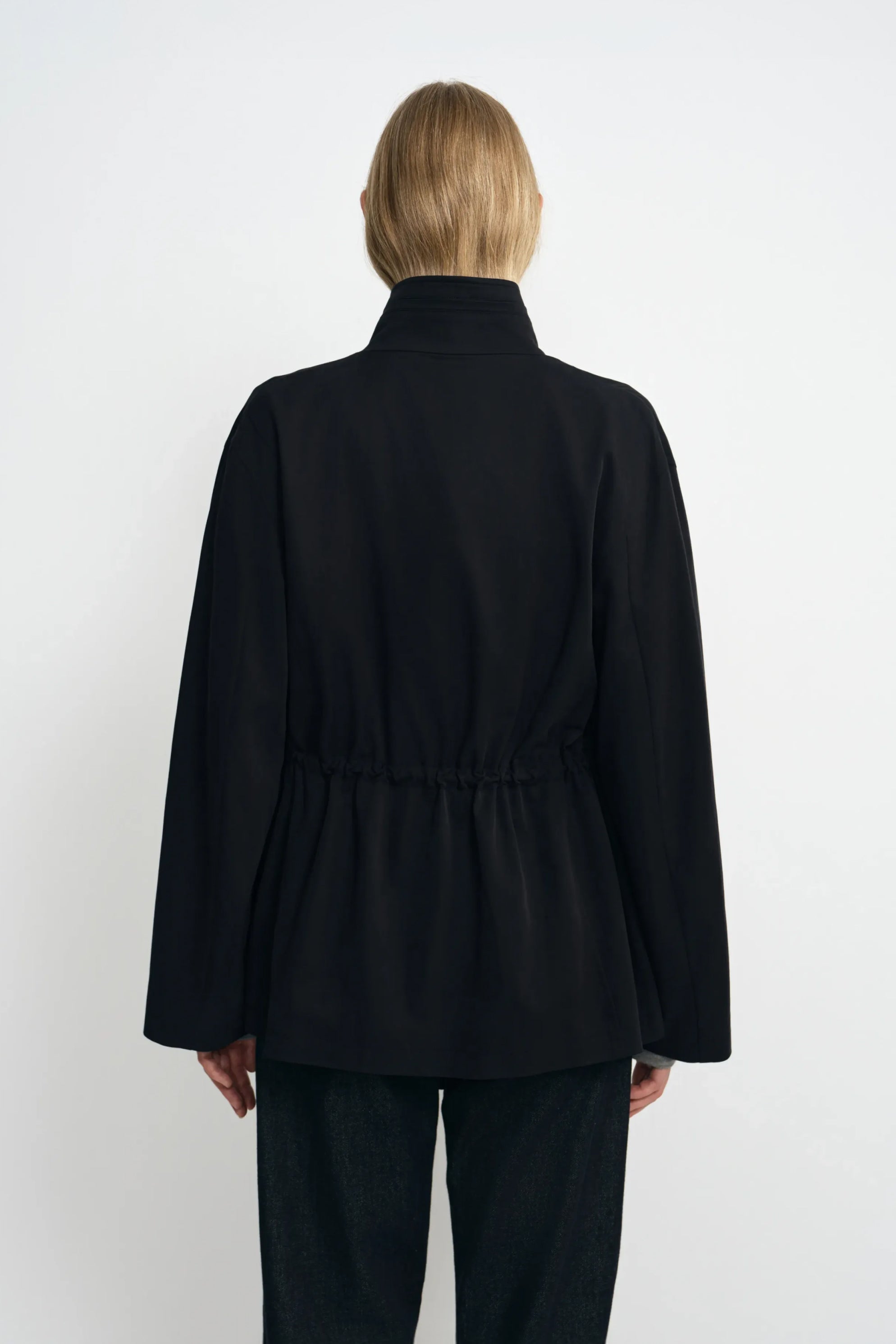 Back view of a model wearing a black jacket, showing the high collar, gathered waist and hem flaring over dark jeans.