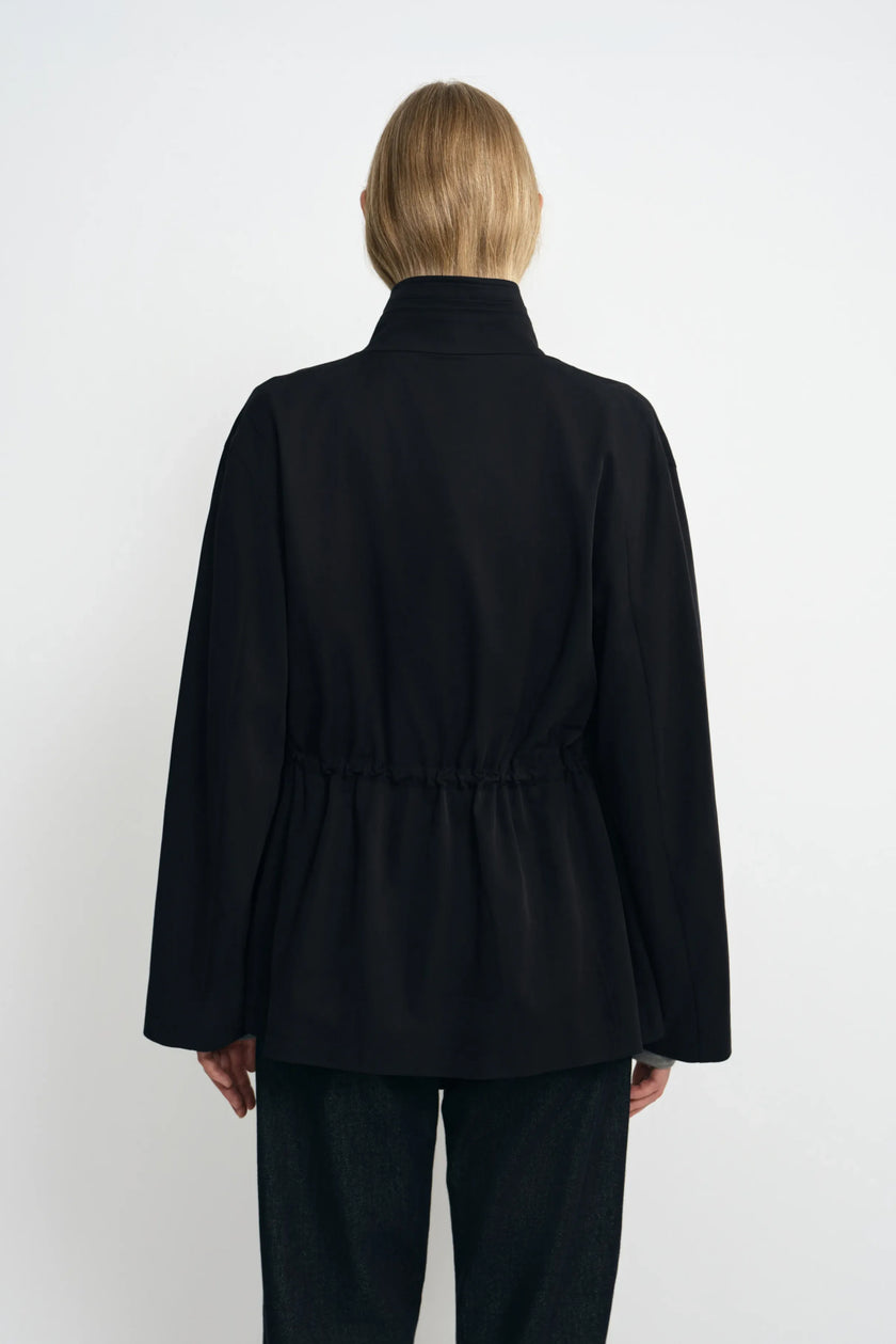 Back view of a model wearing a black jacket, showing the high collar, gathered waist and hem flaring over dark jeans.