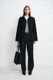 A model wearing a black stand-collar jacket open over grey and white layers, paired with dark jeans, black pointed shoes and a large black tote.