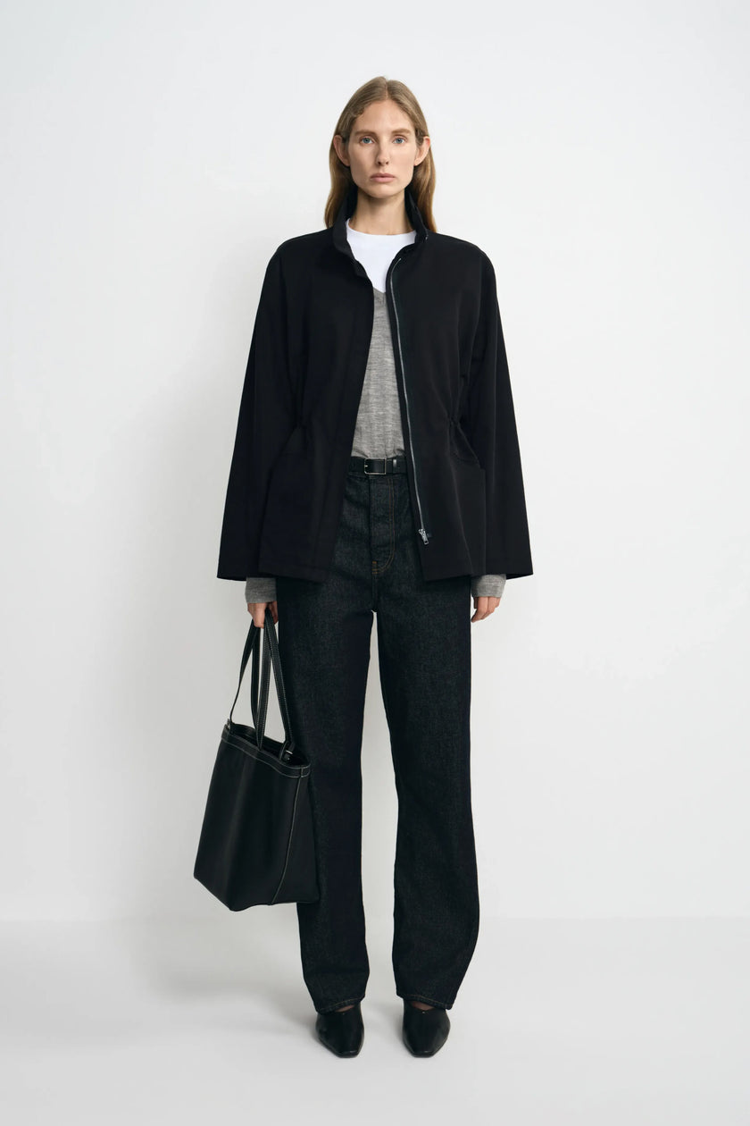 A model wearing a black stand-collar jacket open over grey and white layers, paired with dark jeans, black pointed shoes and a large black tote.