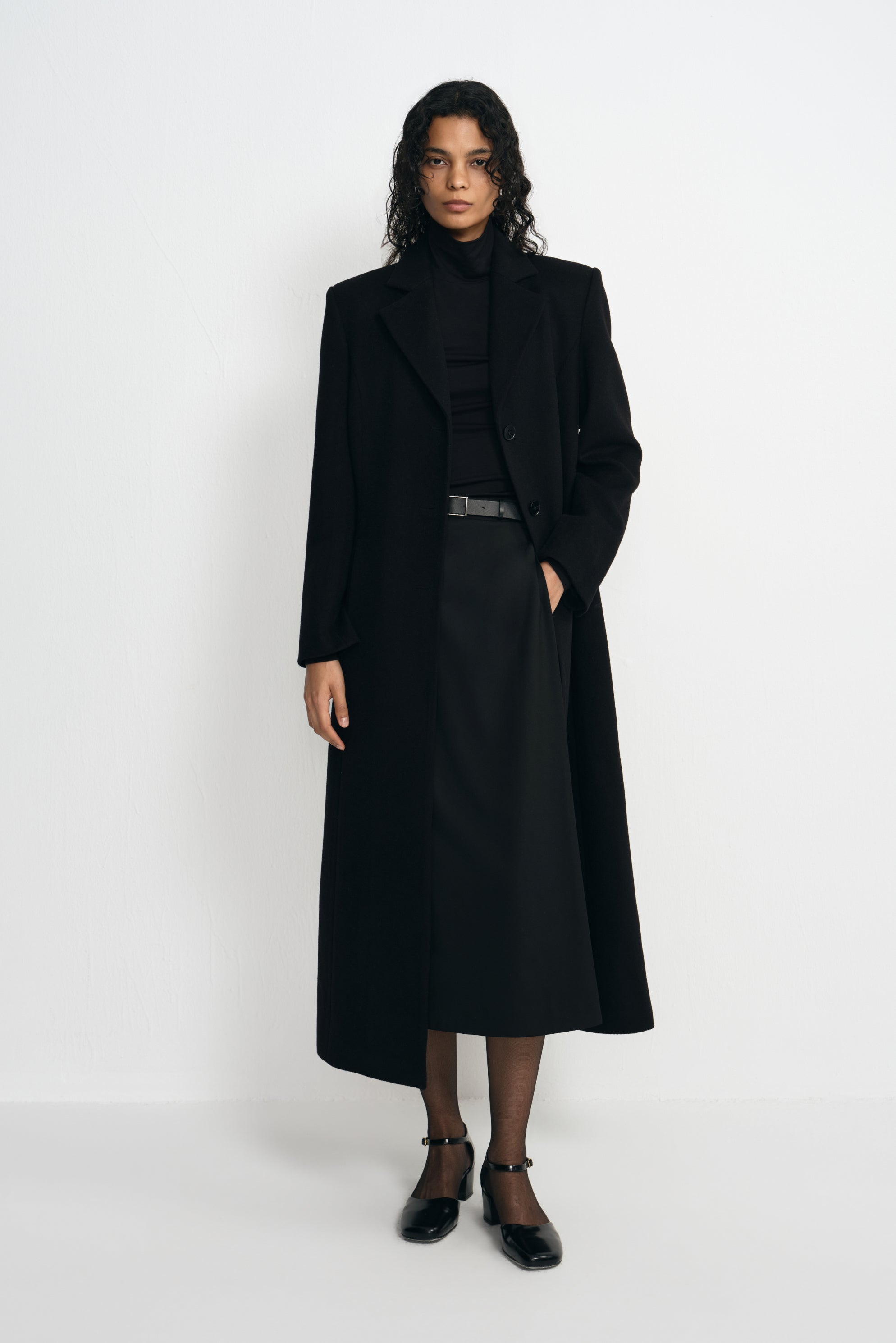 Front view of a model in an open black coat with defined shoulders and a long hem, paired with a black turtleneck, belted midi skirt and ankle-strap shoes.