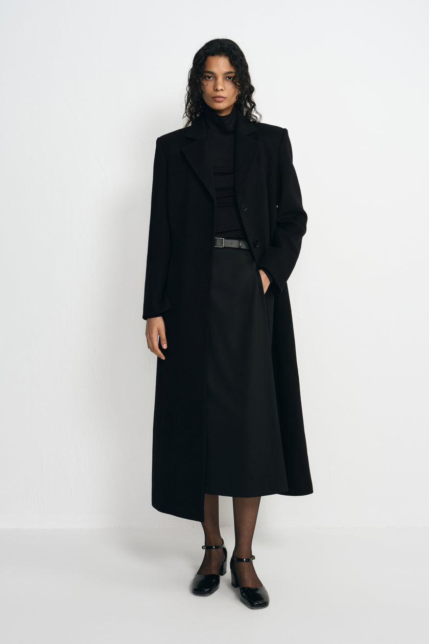 Front view of a model in an open black coat with defined shoulders and a long hem, paired with a black turtleneck, belted midi skirt and ankle-strap shoes.
