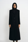 A model facing forward in a buttoned black wool-blend coat, showing sculpted shoulders, notched lapels and a shaped waist over a black turtleneck.