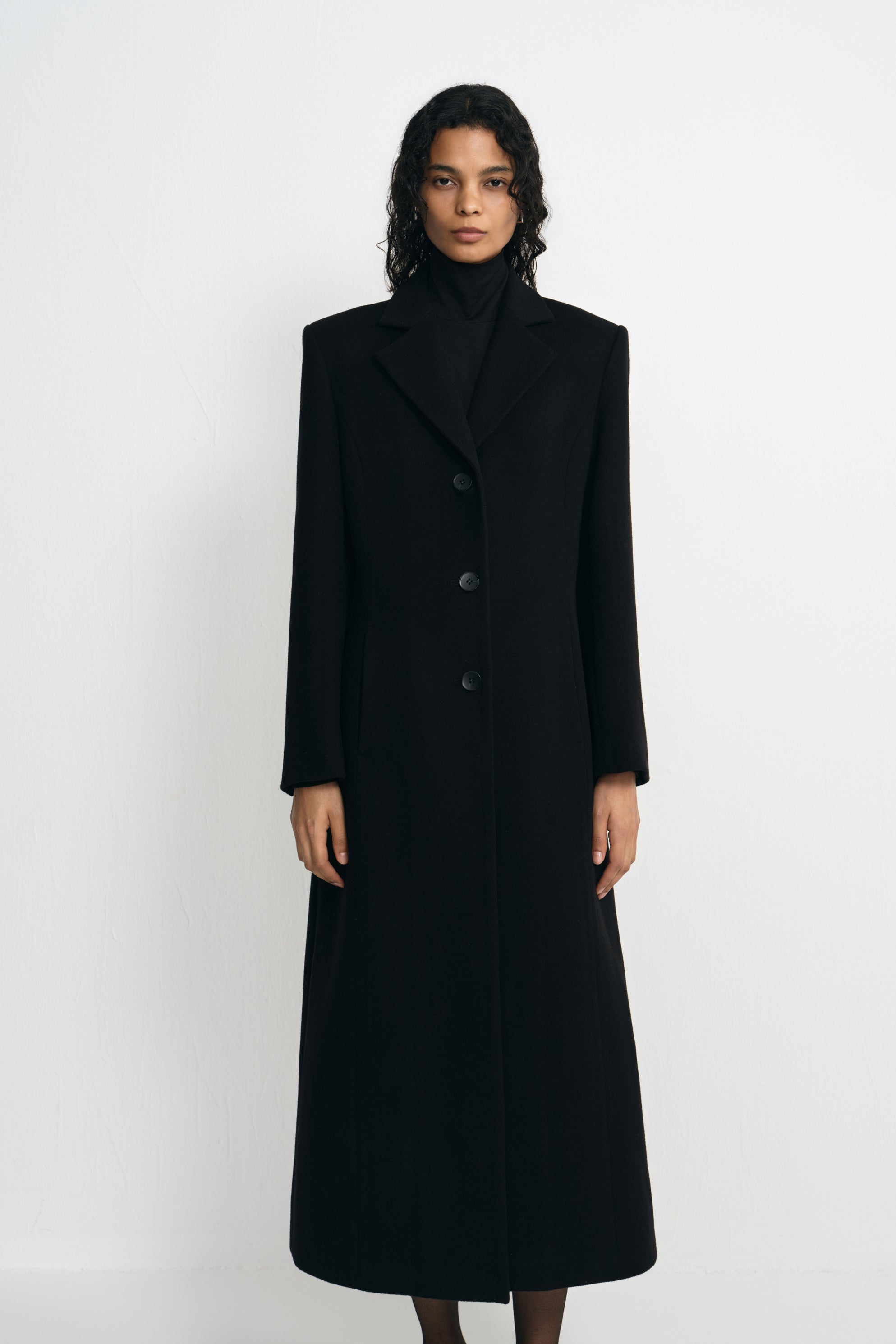 A model facing forward in a buttoned black wool-blend coat, showing sculpted shoulders, notched lapels and a shaped waist over a black turtleneck.