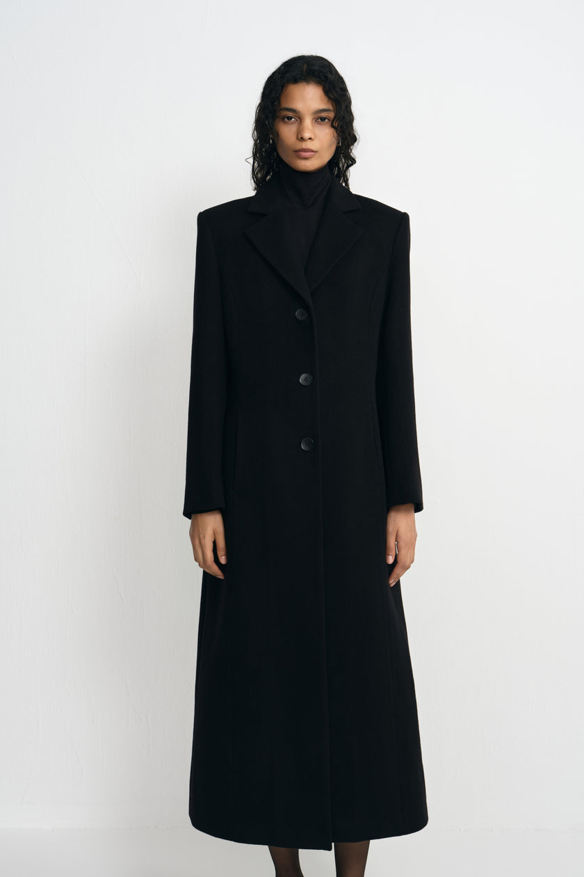 A model facing forward in a buttoned black wool-blend coat, showing sculpted shoulders, notched lapels and a shaped waist over a black turtleneck.
