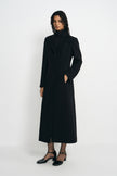 Three-quarter view of a model in a buttoned black coat with defined shoulders and a shaped waist, one hand in a pocket, wearing black ankle-strap shoes.