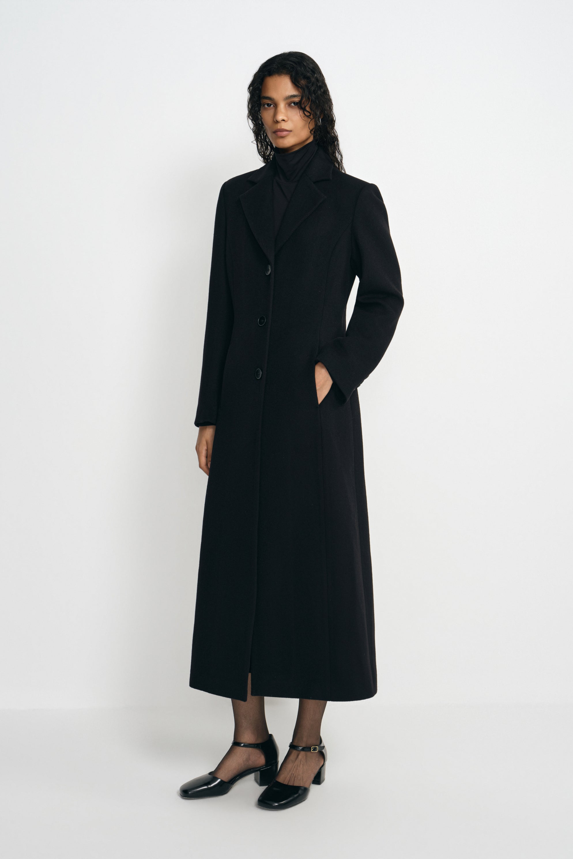 Three-quarter view of a model in a buttoned black coat with defined shoulders and a shaped waist, one hand in a pocket, wearing black ankle-strap shoes.