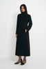 Three-quarter view of a model in a buttoned black coat with defined shoulders and a shaped waist, one hand in a pocket, wearing black ankle-strap shoes.