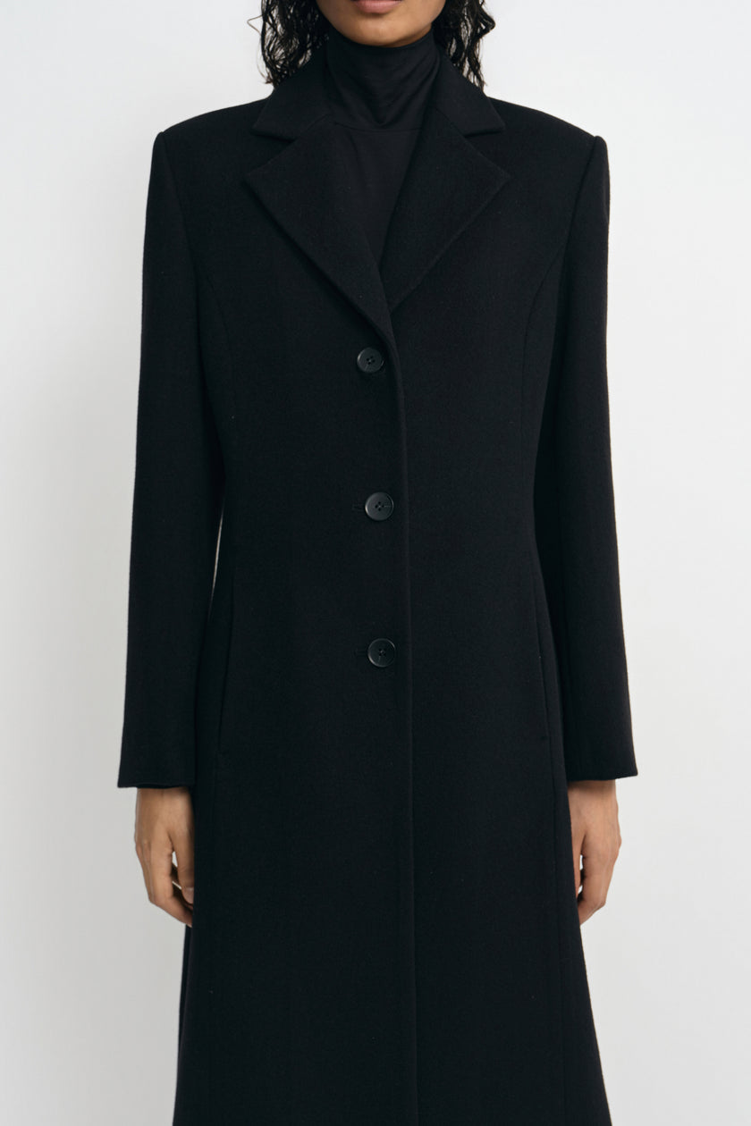 Close-up of a black wool-blend coat on a model, showing the sculpted shoulder line, broad notched lapels and three-button front over a black turtleneck.