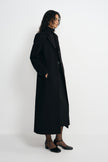 Side view of a model in a long black coat with structured shoulders, worn open over a belted black skirt and paired with black ankle-strap shoes.