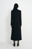 Back view of a model wearing a long black wool-blend coat, showing the defined shoulders and shaped waist above a calf-length hem and black shoes.