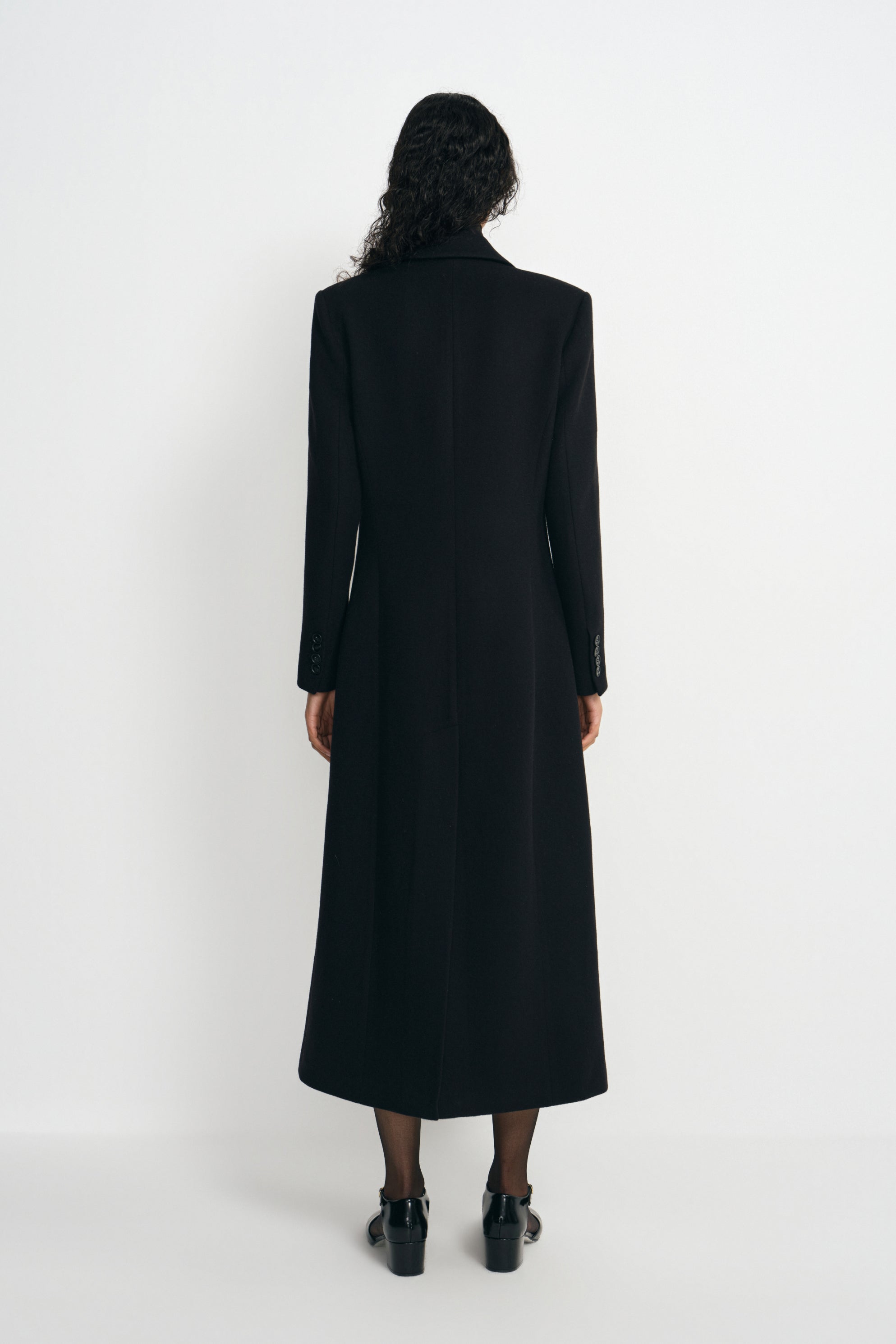 Back view of a model wearing a long black wool-blend coat, showing the defined shoulders and shaped waist above a calf-length hem and black shoes.