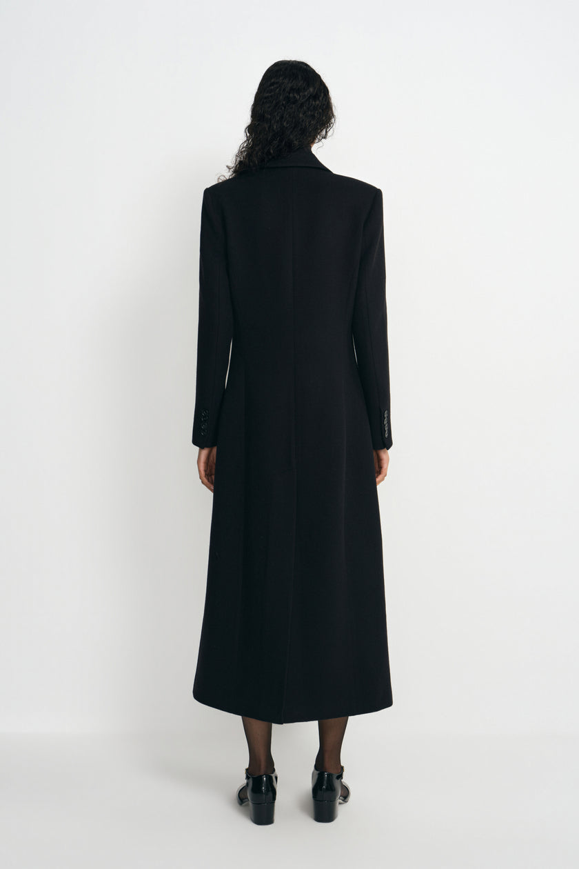 Back view of a model wearing a long black wool-blend coat, showing the defined shoulders and shaped waist above a calf-length hem and black shoes.