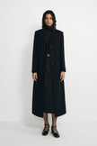 A model wearing a long black wool-blend coat with sculpted shoulders and notched lapels, open over a black turtleneck and belted skirt with ankle-strap shoes.