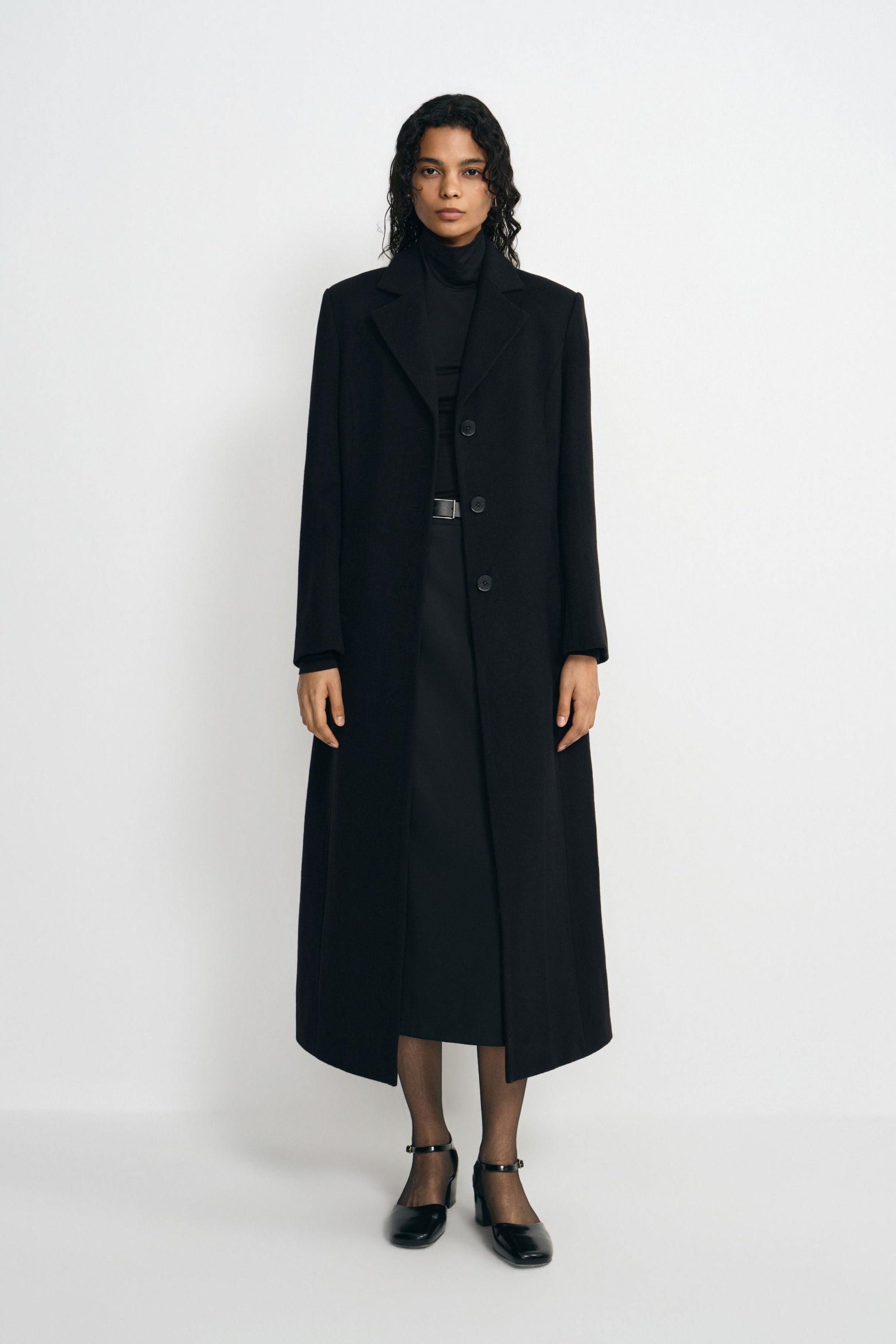 A model wearing a long black wool-blend coat with sculpted shoulders and notched lapels, open over a black turtleneck and belted skirt with ankle-strap shoes.