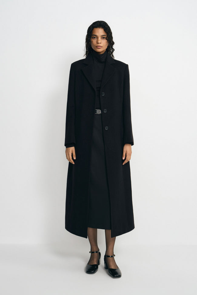 A model wearing a long black wool-blend coat with sculpted shoulders and notched lapels, open over a black turtleneck and belted skirt with ankle-strap shoes.