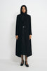 A model wearing a long black wool-blend coat with sculpted shoulders and notched lapels, open over a black turtleneck and belted skirt with ankle-strap shoes.