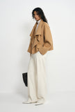 Side view of a model in a tan-beige short coat, showing the layered waterfall collar and broad sleeves, styled with cream trousers and pointed white shoes.