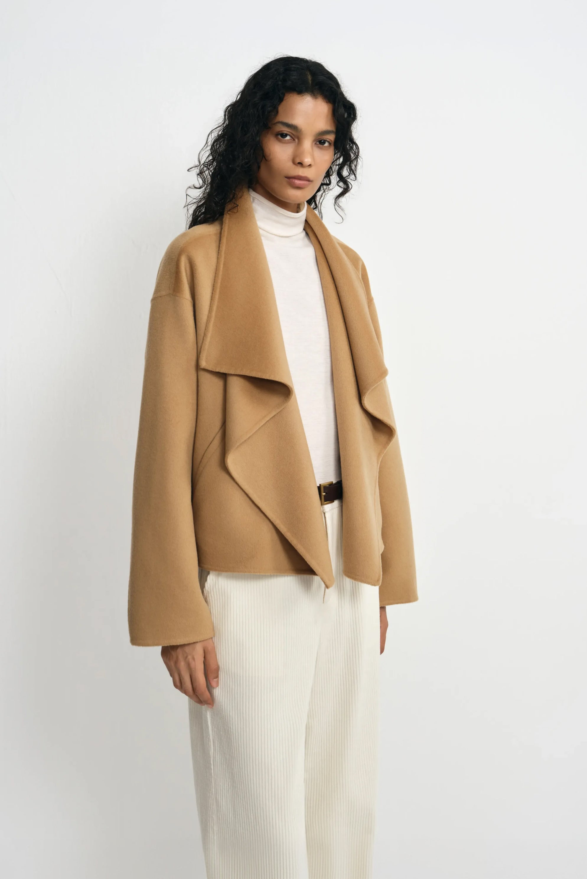 Three-quarter close-up of a model in a tan-beige coat with a folded waterfall collar, worn over a cream turtleneck and trousers with a narrow dark belt.