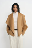 Front detail of a tan-beige coat on a model, with the collar spread into broad points over a cream turtleneck and belted cream trousers.