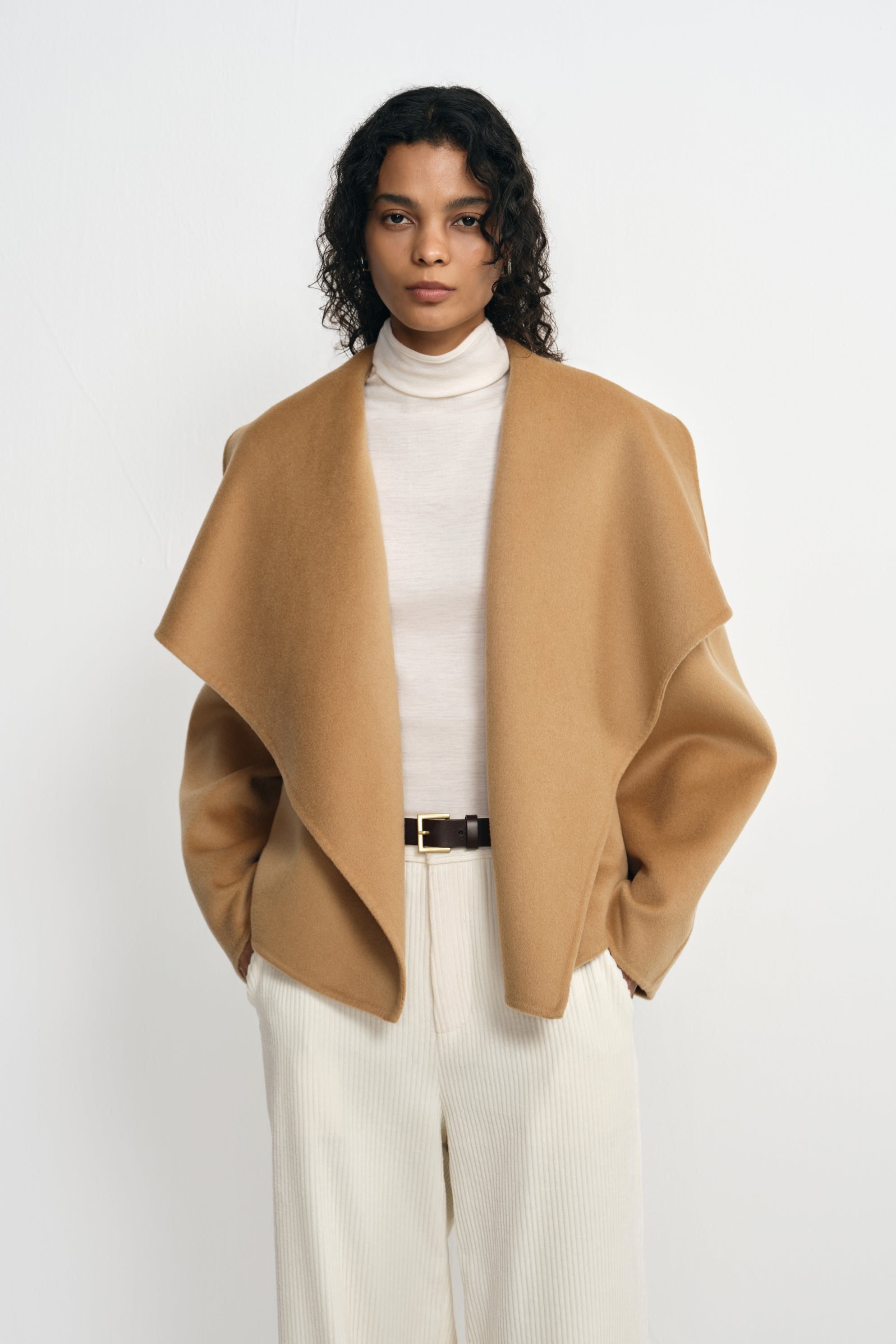 Front detail of a tan-beige coat on a model, with the collar spread into broad points over a cream turtleneck and belted cream trousers.