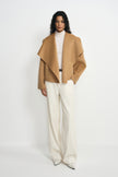 A model wearing a tan-beige short coat with the collar opened wide, styled with a cream turtleneck, wide-leg cream trousers and pointed white shoes.