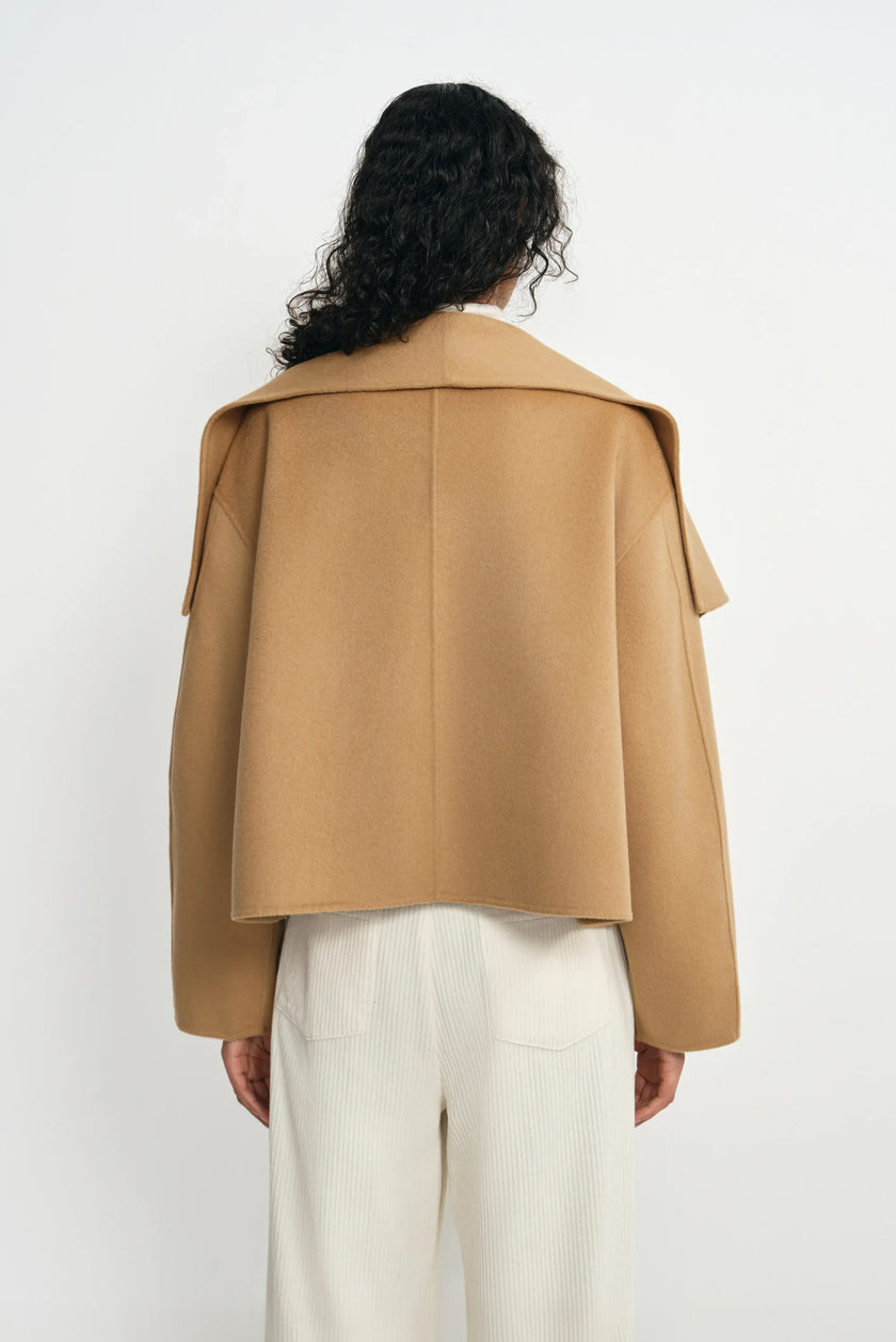 Back view of a tan-beige wool-cashmere short coat on a model, showing the wide collar across the shoulders, center seam and straight hem above cream trousers.