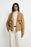 A model facing forward in a tan-beige waterfall coat, showing the cascading collar folds over a cream turtleneck and trousers with a slim dark belt.