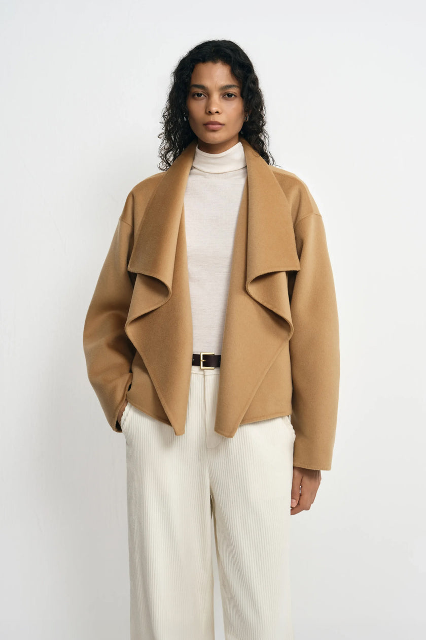 A model facing forward in a tan-beige waterfall coat, showing the cascading collar folds over a cream turtleneck and trousers with a slim dark belt.