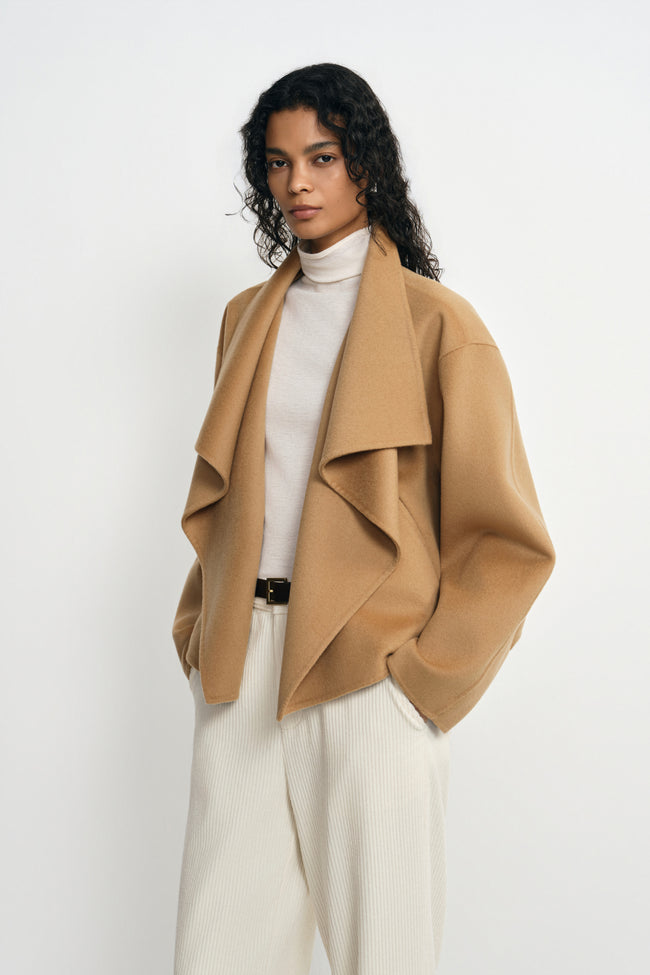 Angled close-up of a model wearing a tan-beige wool-cashmere coat, showing the layered waterfall collar and full sleeve beside cream belted trousers.