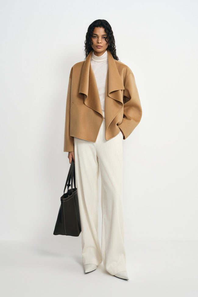 A model wearing a tan-beige wool-cashmere short coat with a cascading waterfall collar, paired with a cream turtleneck, wide-leg trousers and white shoes.