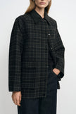 Close-up of a grey plaid jacket on a model, showing the black pointed collar, metal snaps and rectangular flap pocket above dark belted jeans.