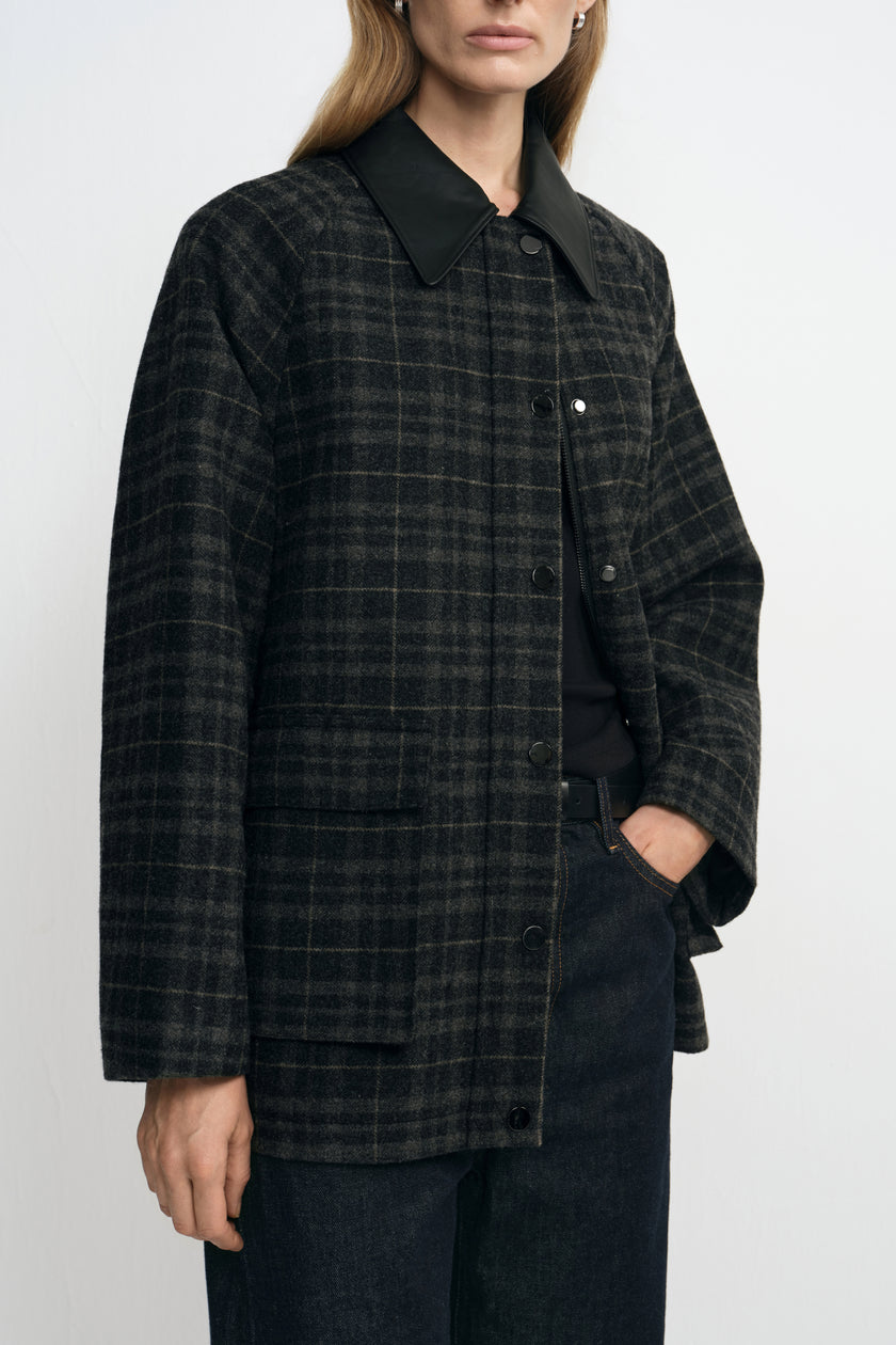 Close-up of a grey plaid jacket on a model, showing the black pointed collar, metal snaps and rectangular flap pocket above dark belted jeans.