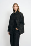 Three-quarter view of a model wearing a grey plaid jacket with a black collar and wide sleeves, layered over a black top and dark jeans.