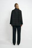 Back view of a model in a grey plaid jacket, showing the checked pattern across its straight back and hem, paired with dark jeans and black shoes.