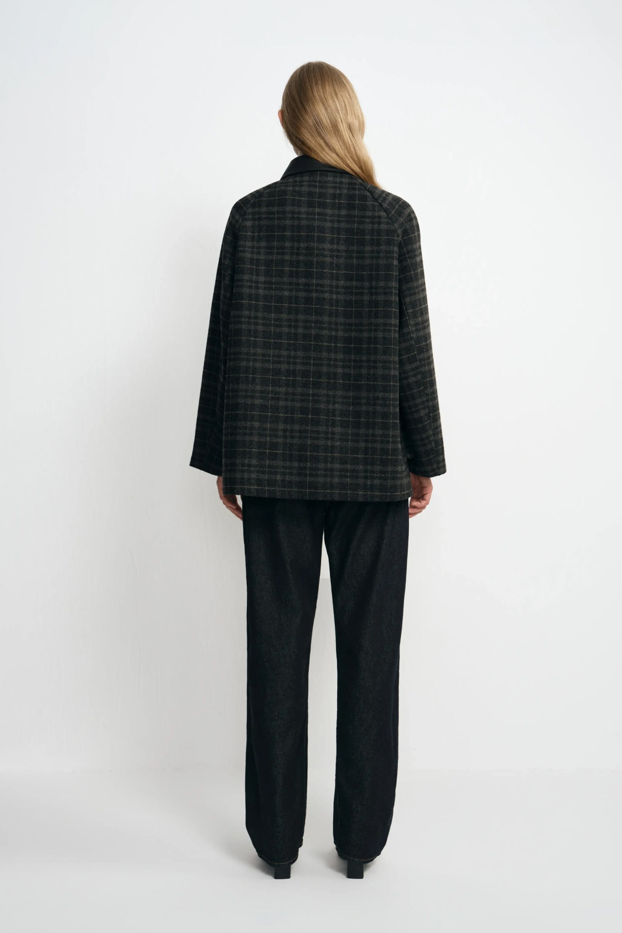 Back view of a model in a grey plaid jacket, showing the checked pattern across its straight back and hem, paired with dark jeans and black shoes.