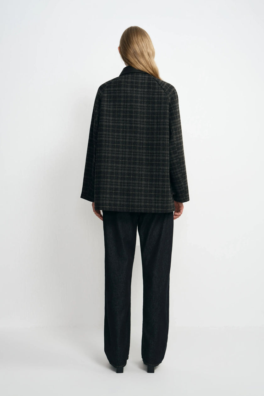 Back view of a model in a grey plaid jacket, showing the checked pattern across its straight back and hem, paired with dark jeans and black shoes.