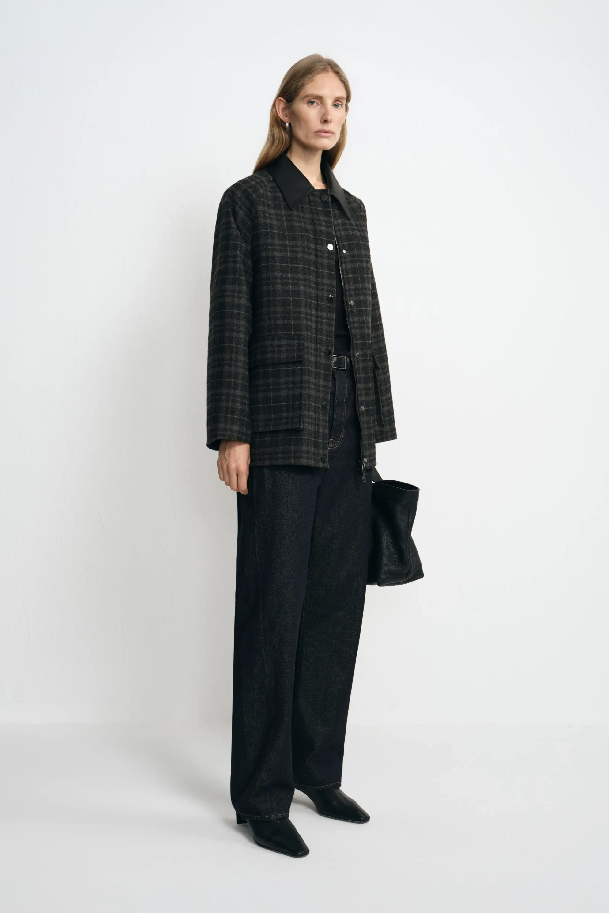 Angled view of a model in a grey plaid jacket with a contrasting black collar, styled with a black top, dark jeans, pointed shoes and a black handbag.