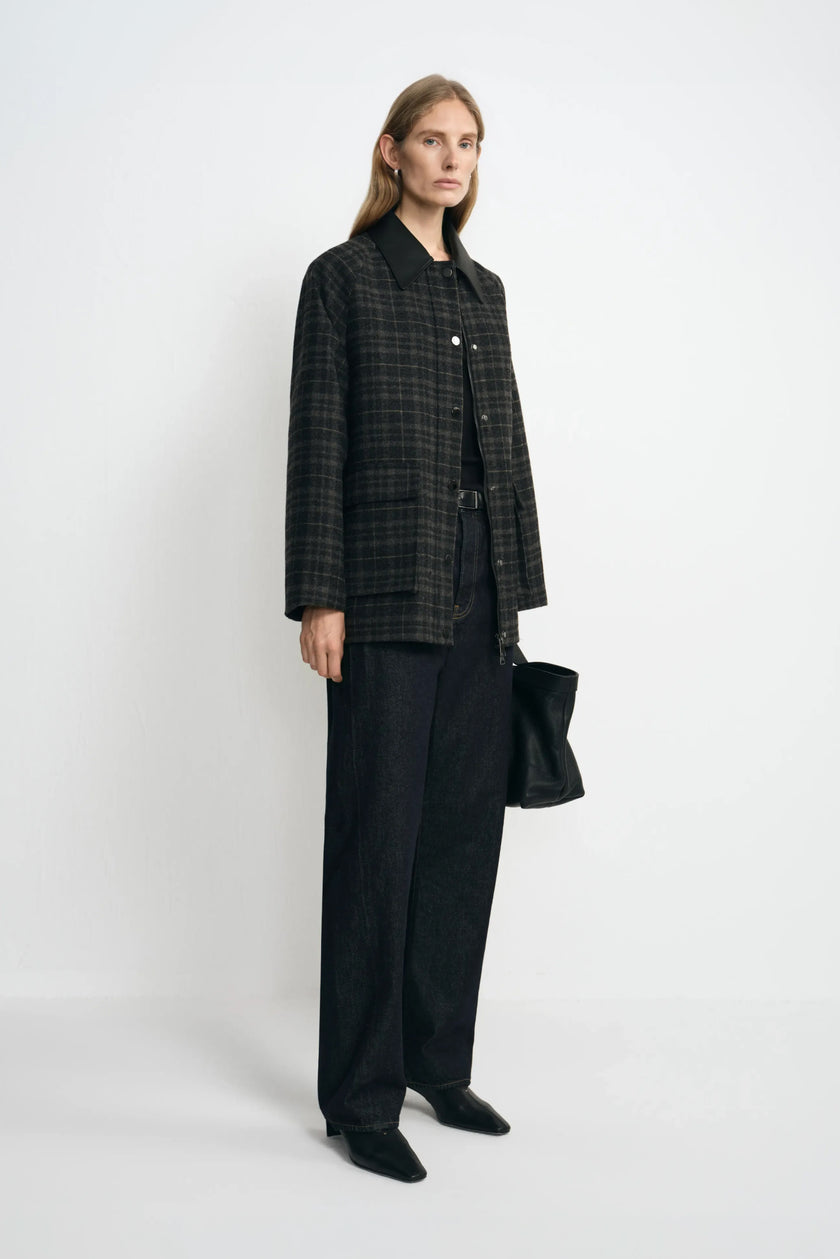 Angled view of a model in a grey plaid jacket with a contrasting black collar, styled with a black top, dark jeans, pointed shoes and a black handbag.