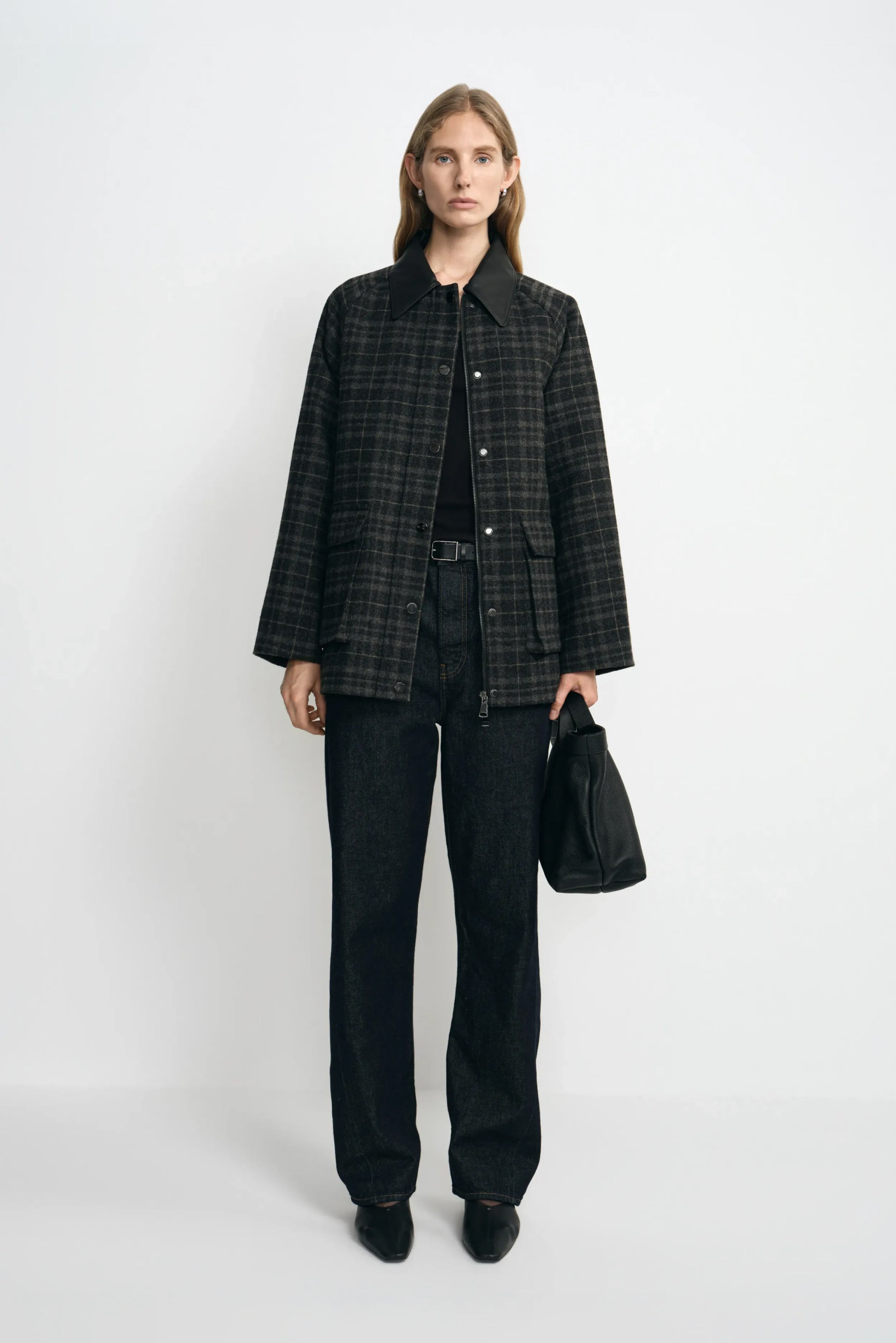 A model facing forward in a grey plaid wool-blend jacket with a black sheepskin collar, paired with dark jeans, black pointed shoes and a black bag.