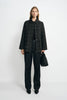 A model facing forward in a grey plaid wool-blend jacket with a black sheepskin collar, paired with dark jeans, black pointed shoes and a black bag.